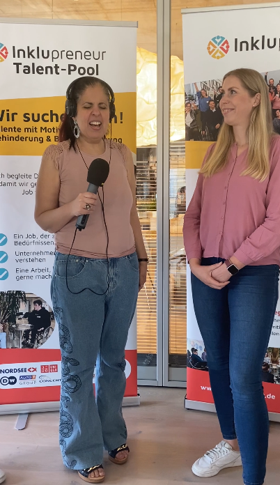 Eine Mitarbeitende von Nordsee, Cornelia Kraatz steht mit Amy Zayed von Inklupreneur in einer Interviewsituation nebeneinander. Im Hintergrund steht ein Aufsteller zum Inklupreneur Talentpool. Die Aufmerksamkeit liegt bei Amy.