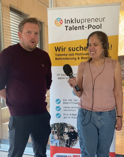 Ein Mitarbeitender von Nordsee, Sebastian Pape steht mit Amy Zayed von Inklupreneur in einer Interviewsituation nebeneinander. Im Hintergrund steht ein Aufsteller zum Inklupreneur Talentpool. Die Aufmerksamkeit liegt bei Amy.