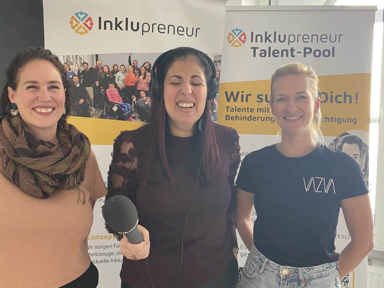 Amy Zayed von Inklupreneur steht im Zentrum des Bildes. Sie hat Kopfhörer auf, hält ein Mikrophon in der Hand und lacht in die Kamera. Im Hintergrund steht ein Aufsteller mit dem Schriftzug "Inklupreneur". Am Bildrand auf beiden Seiten sind Christin Kannenberg und  Franziska Weinhold beide halb abgebildet.